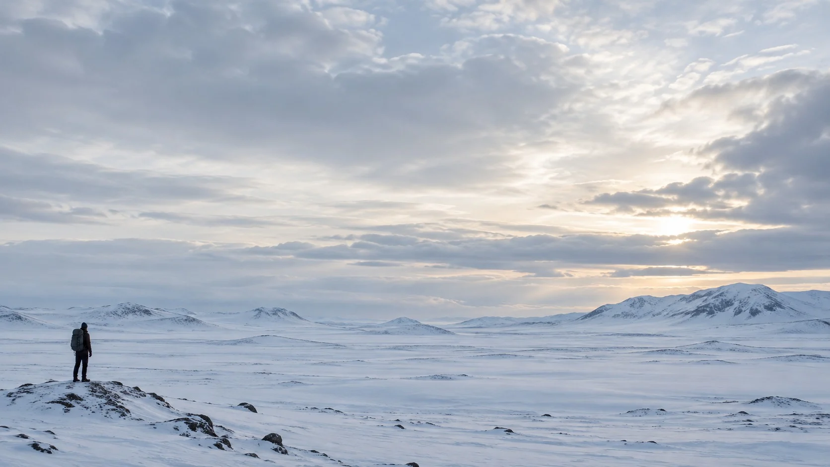A single hiker stands very small in the lower corner of a vast, empty snowfield, with most of the frame left open to sky and snow