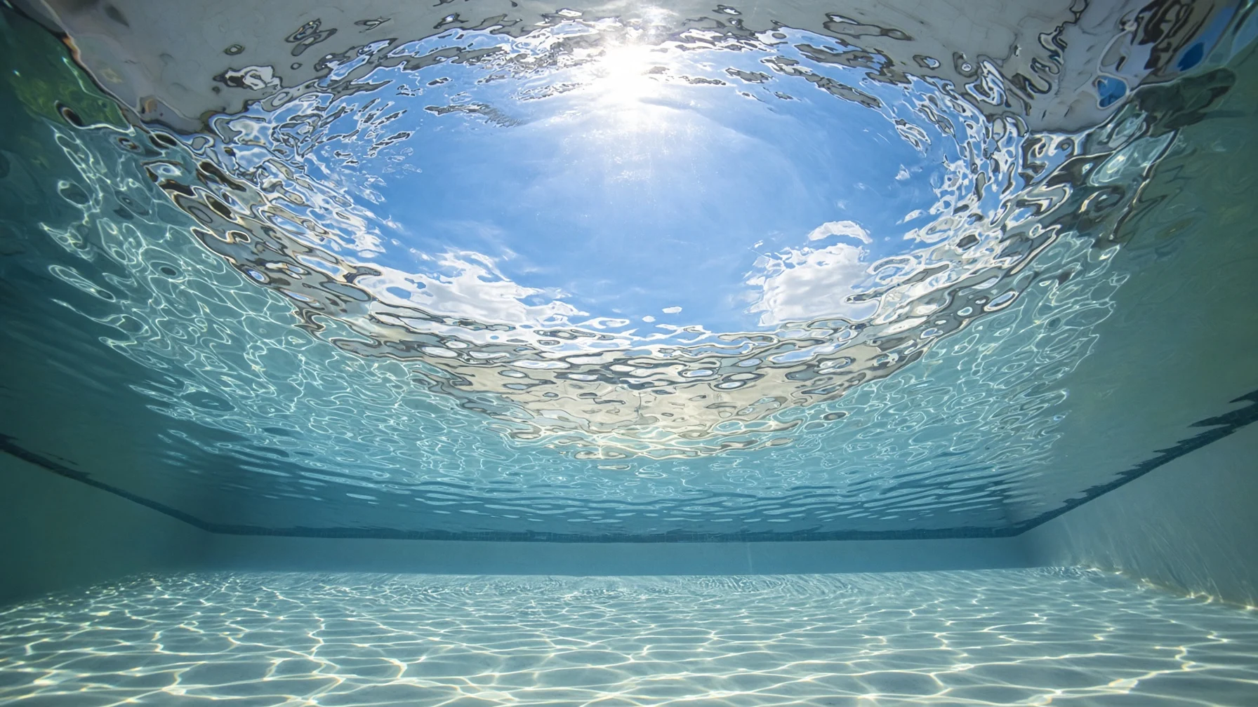 晴れた日の屋外プールの底から見上げた水面に、青い空と日差しが丸い窓のように見え、その外側の水面が鏡のようにプールの底を映している様子